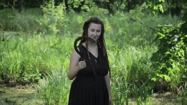 Una giovane ragazza affascinante con dreadlocks posa per la fotocamera agitando una falce e sorridendo furbescamente sullo sfondo degli alberi. Primo piano — Video Stock