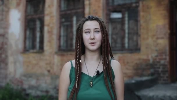 Ein nettes junges Mädchen mit Dreadlocks sagt vor dem Hintergrund eines alten Backsteinhauses leidenschaftlich etwas in ein Mikrofon, das an der Kleidung befestigt ist. Nahaufnahme — Stockvideo