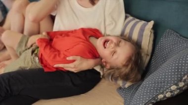 Anne, küçük oğlunu kucağında tutar ve onu gıdıklarken kızı da babasıyla odada kanepede oturur. Ağır çekim. Yakın plan.