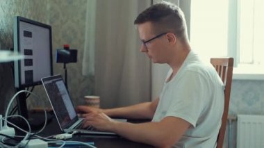 Odaklanmış genç bir erkek serbest yazar odasındaki bir masada bilgisayar üzerinde çalışıyor. Yandan bak. Yakın plan.