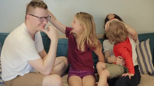 Una familia joven y feliz con dos hijos está sentada en el sofá de la habitación y riéndose entre sí con el pelo. Primer plano — Vídeo de stock