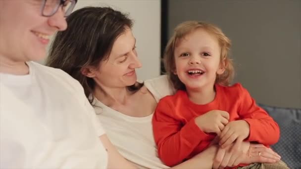 Un bambino in t-shirt rossa e capelli biondi siede tra le braccia della madre sul divano accanto al padre e cerca di leccare la madre. La famiglia ride insieme. Primo piano — Video Stock