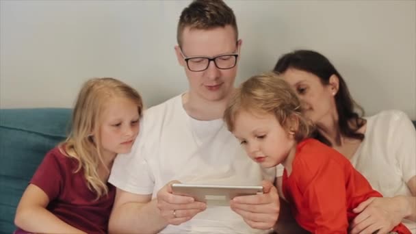 Una famiglia felice con due bambini si siede sul divano e guarda il tablet insieme. Il piccolo figlio fa capolino in modo interessante allo schermo. Primo piano — Video Stock