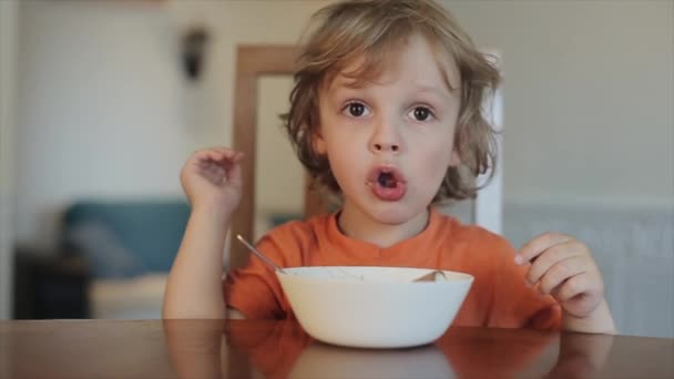Divertido niño come helado en la mesa de la cocina y señala con el dedo algo detrás. En cámara lenta. Primer plano — Vídeos de Stock
