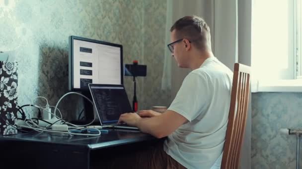 Молодий чоловік працює вдома за столом відразу на двох комп'ютерах, ретельно вивчаючи інформацію з одного і друкуючи з іншого. Вид збоку. Крупним планом — стокове відео