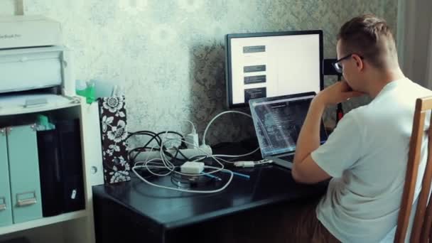 Un joven hombre freelance trabaja enfocado en sentarse en una mesa en una habitación estudiando cuidadosamente lo dado con dos computadoras. Vista desde atrás. Primer plano — Vídeos de Stock