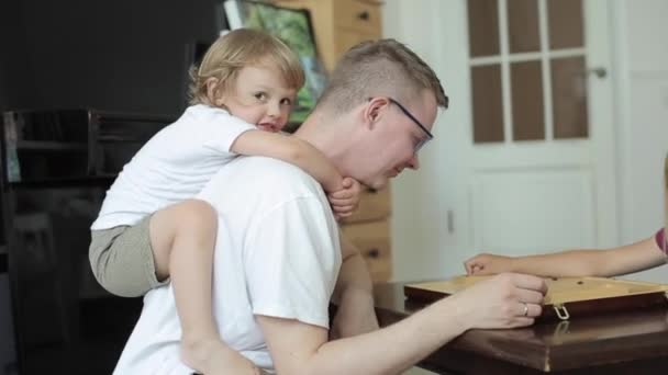En liten rastlös pojke sitter runt sina fäders hals medan han leker med sin dotter i brädspel sitter vid ett bord i rummet. Utsikt från sidan. Närbild — Stockvideo