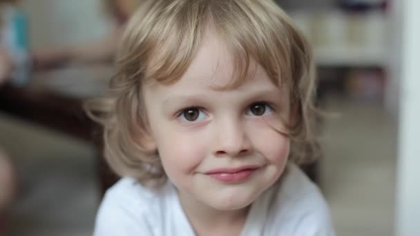 Porträt eines charmant lächelnden blonden Jungen mit lockigem Haar und großen braunen Augen in einem weißen T-Shirt. Nahaufnahme — Stockvideo