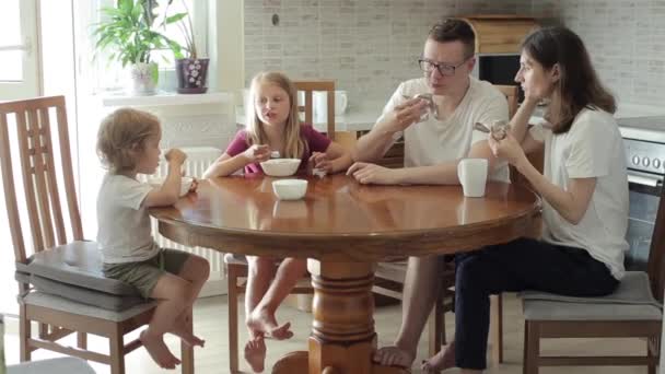 Una giovane famiglia felice con un figlio piccolo e una figlia maggiore si siede a un grande tavolo rotondo in cucina a piedi nudi mangiando gelato e parlando. Primo piano — Video Stock
