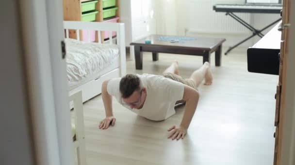 Un joven atractivo con gafas y una camiseta blanca entrena en casa empujando desde el suelo y saltando con las manos en alto. Primer plano — Vídeos de Stock