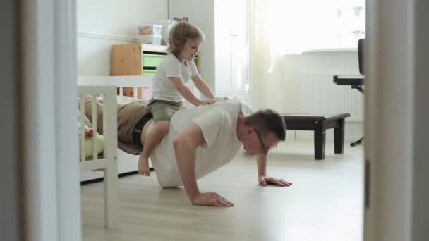 Un niño rubio con una camiseta blanca se sienta en la espalda de su padre fuerte y sonríe felizmente mientras empuja hacia arriba desde el suelo de la habitación. Primer plano — Vídeos de Stock