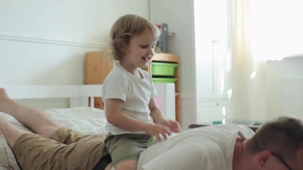 A little blond boy with big brown eyes sits on his fathers back imagining that this is a horse while his father does an exercise and push-up off the floor. Close-up — Stock Video