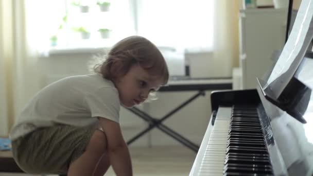 Een blond jongetje met krullen en grote bruine ogen klimt op een stoel en drukt zachtjes op de pianotoetsen. Close-up — Stockvideo