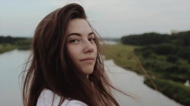 Büyüleyici genç bir kız nehir ve ormanın arka planına karşı köprüde yürüyor ve doğrudan kameraya bakıyor. Yavaş çekim. Yakın plan.