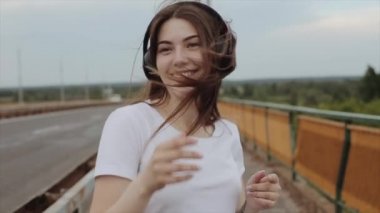 Neşeli genç bir kız bir yaz günü yol köprüsünde yürürken büyük kulaklıklarla müzik dinleyerek dans eder. Ağır çekim. Yakın plan.