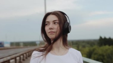 Bir yaz günü köprüde yürüyen ve büyük siyah kulaklıklarla müzik dinleyen genç ve çekici bir kızın portresi. Yavaş çekim