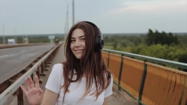 Veselá mladá dívka stojí na můstku ve velkých černých sluchátkách a tancích a poslouchá hudbu. Zpomal. Detailní záběr — Stock video