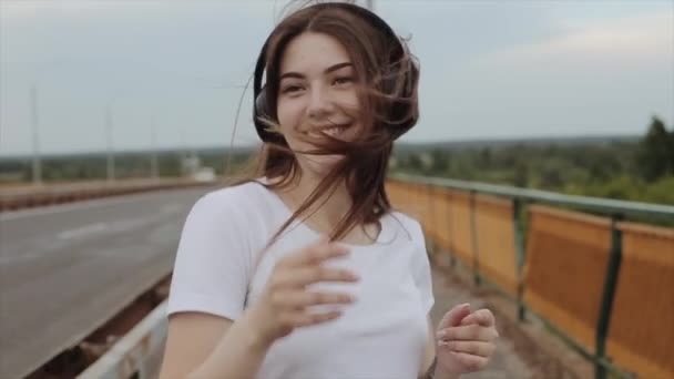 Uma jovem alegre dança ouvindo música em grandes fones de ouvido andando na ponte da estrada em um dia de verão. Movimento lento. Close-up — Vídeo de Stock