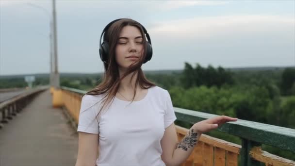 Romantisches junges Mädchen läuft auf der Brücke im Hintergrund der Natur und hört Musik mit schwarzen Kopfhörern. Zeitlupe. Nahaufnahme — Stockvideo