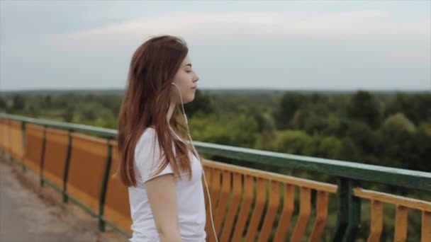 En romantisk ung flicka går över bron i hörlurar för en mobiltelefon och njuter av utsikten över naturen och musiken. Sakta i backarna. Närbild — Stockvideo
