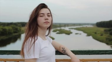Kolunda dövmesi olan şehvetli genç bir kız köprünün üzerinde doğanın arka planına doğru yürüyor ve uzun saçlarını düzeltiyor. Yavaş çekim. Yakın plan.