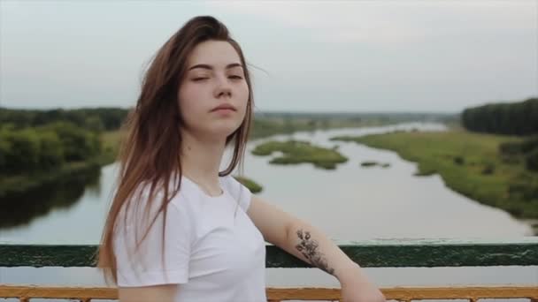 Chica joven sensual con un tatuaje en el brazo camina sobre el puente contra el telón de fondo de la naturaleza y corrige su cabello largo. En cámara lenta. Primer plano — Vídeo de stock