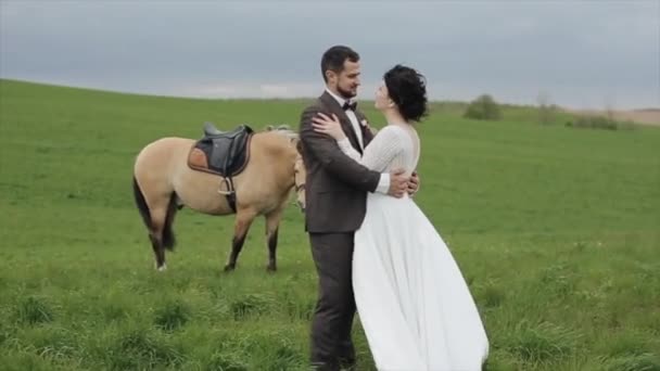 Счастливые молодожены целуются на прогулке по ранчо рядом с лошадью, которая ест траву на лугу. Крупный план — стоковое видео