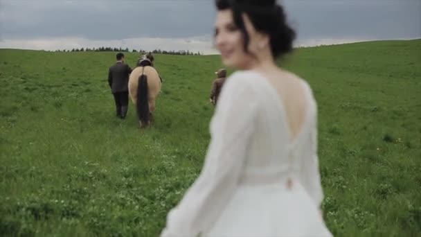 Recién casados en un paseo por un rancho. El novio es guiado por un caballo. La novia camina por detrás y mira en cámara sonriendo. La cámara cambia de enfoque. Primer plano — Vídeos de Stock