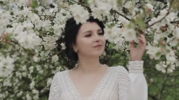 Een charmante jonge vrouw met een prachtig kapsel en een witte kanten jurk staat tussen de bloeiende appelbomen. Een close-up. Camera verandert focus — Stockvideo