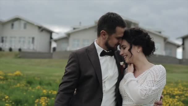 Glada nygifta går genom den blommande ängen mot bakgrund av vackra stugor. Brudgummen kysser bruden i pannan. Närbild. Långsamma rörelser — Stockvideo