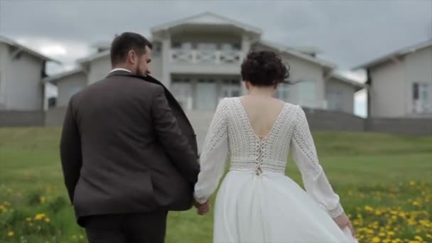 Η νύφη και ο γαμπρός σε μια βόλτα μέσα από το ανθισμένο λιβάδι σε μια θυελλώδη καλοκαιρινή μέρα. Θέα από πίσω. Κοντινό πλάνο. Η κάμερα μετακινείται από πάνω προς τα κάτω — Αρχείο Βίντεο