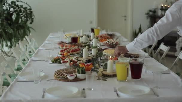 O garçom acende velas em vasos de vidro em uma mesa festivamente decorada com muitos pratos com alimentos e lanches diferentes. Close-up — Vídeo de Stock