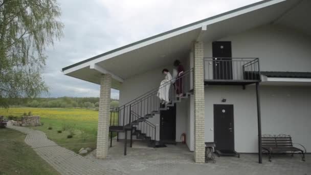 La novia desciende cuidadosamente los escalones de hierro de la casa de campo. La dama de honor la ayuda y lleva el velo. Boda en una casa de campo — Vídeos de Stock