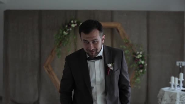 A funny silly stupid young bearded groom in a suit fooling around and walks around the room for the wedding ceremony and jokes with a videographer showing finger gestures to the camera and laughing — Stock Video