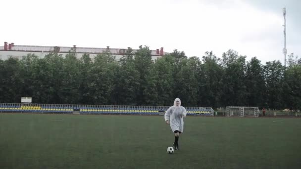 Un futbolista en un impermeable hace ejercicios con la pelota bajo la lluvia mientras su equipo resuelve los momentos de juego en la otra mitad del campo — Vídeos de Stock