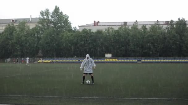 De camera richt zich op de regendruppels op de wazige achtergrond van de voetballer in een regenjas met de bal op het veld en verandert dan van focus op de speler — Stockvideo