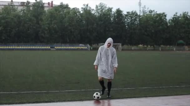 Un jugador del club de fútbol con un impermeable blanco patea la pelota caminando en cámara y sonríe bajo la lluvia. Primer plano — Vídeos de Stock