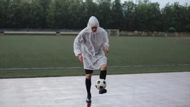 Beyaz yağmurluk giyen bir futbolcu, yağmur altında stadyum koşu bandında serbest stil bir futbol topu gösteriyor. Yakın plan. — Stok video