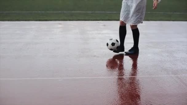 Ένας ποδοσφαιριστής με ένα λευκό αδιάβροχο με ένα ασυνήθιστο κούρεμα στη βροχή γεμίζει την μπάλα στη συνέχεια αφήνει την μπάλα και τα φύλλα κοιτάζοντας πίσω στην κάμερα. Κοντινό πλάνο — Αρχείο Βίντεο