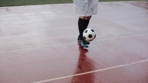 Yağmurda alışılmadık bir saç kesimi olan beyaz yağmurluk giyen bir futbolcu topu doldurur ve topu bırakıp kameraya bakar. Yakın plan. — Stok video