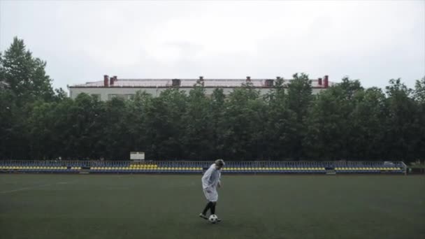 Гравець футбольної команди в плащі тренується на полі з м'ячем під дощем. Камера змінює фокус від футболіста до крапель — стокове відео