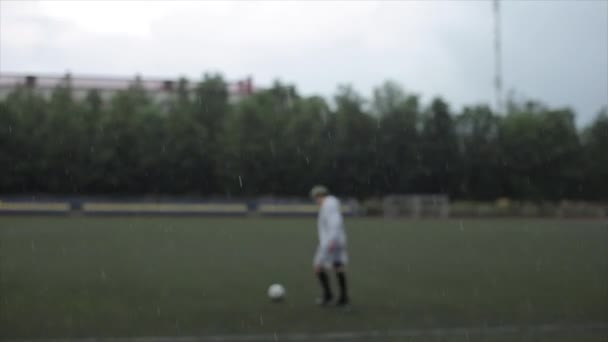 Hráč fotbalového týmu v pláštěnce trénuje na hřišti s míčem v dešti. Kamera mění zaměření z fotbalisty na kapky deště — Stock video
