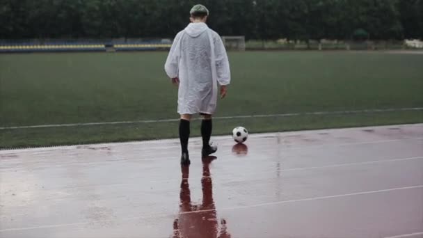 Ένας ποδοσφαιριστής με ένα λευκό αδιάβροχο στην προπόνηση κλωτσάει την μπάλα στο γήπεδο ποδοσφαίρου στη βροχή. Προβολή από πίσω — Αρχείο Βίντεο