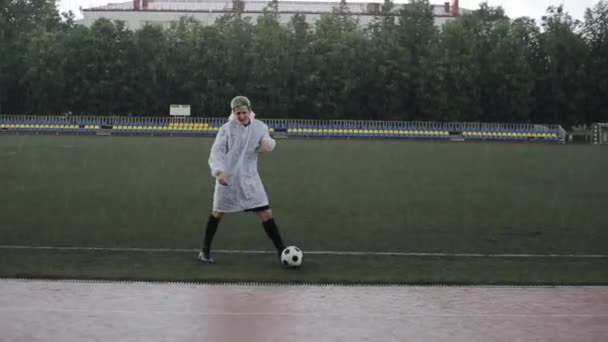 Beyaz yağmurluk giyen bir futbolcu, yağmur altında stadyum koşu bandında serbest stil bir futbol topu gösteriyor. Yakın plan. — Stok video