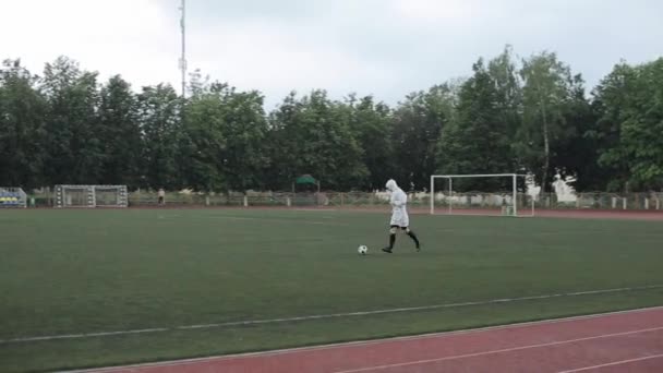 Beyaz yağmurluk giyen bir futbolcu, yağmur altında stadyum koşu bandında serbest stil bir futbol topu gösteriyor. Yakın plan. — Stok video