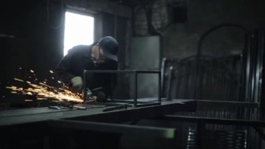 Atölyede siyah üniformalı, koruyucu gözlüklü ve eldivenli bir adam öğütme makinesiyle metal keser. Metalden kıvılcımlar çıkar. Yan görünüm