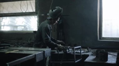 Kaynak miğferi ve özel giysiler içindeki çilingir atölyenin masasına metal bir yapı asıyor. Yan görünüm