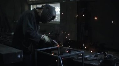 Kaynak miğferi ve özel giysiler içindeki çilingir atölyenin masasına metal bir yapı asıyor. Yan görünüm