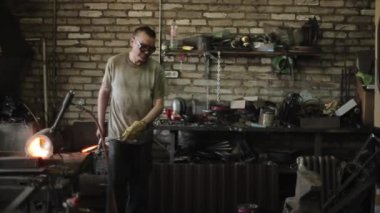 Demirci, demircilerin ocak manzarasının yanında duran örsün üzerinde kızgın demir bir çubuğa vurur. Yakın plan.