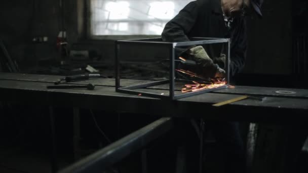 Um homem de uniforme preto em óculos de proteção e luvas na oficina corta metal com uma máquina de moer. Faíscas voam de metal de. Vista lateral — Vídeo de Stock